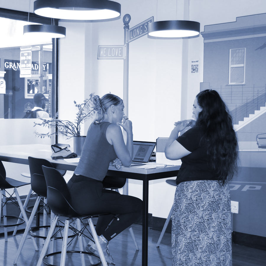 Modern coffee shop scene with two women engaging in conversation and working on laptops.